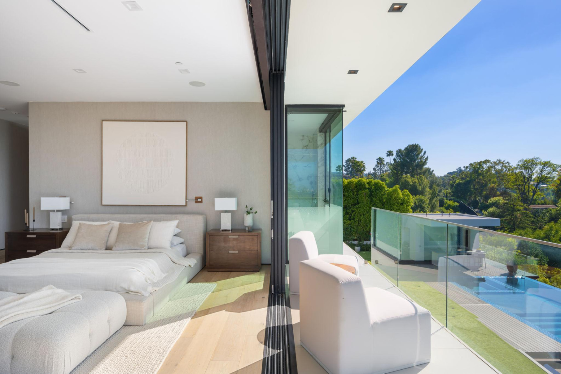 Light-filled bedroom opening onto a glass balcony overlooking a pool and treetops